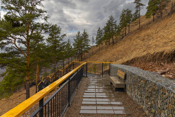 Obraz premium Empty tourist trail in rainy springtime day in the mountain hillside. Metal staircase with wooden railing among pine trees on Torgashinsky ridge in Krasnoyarsk, Russia
