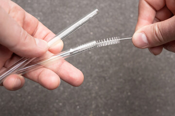Hands holding reusable glass straws for cocktails and cleaning them with a special brush on a blurred gray background. Ecological concept. 