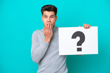 Young handsome caucasian man isolated on blue bakcground holding a placard with question mark symbol with surprised expression