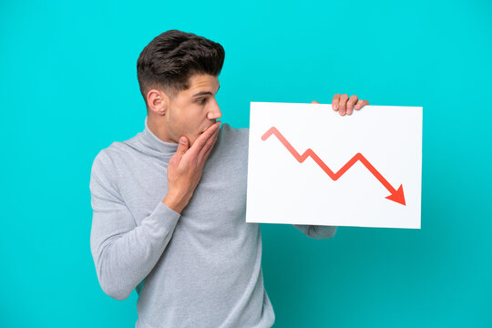 Young handsome caucasian man isolated on blue bakcground holding a sign with a decreasing statistics arrow symbol with surprised expression