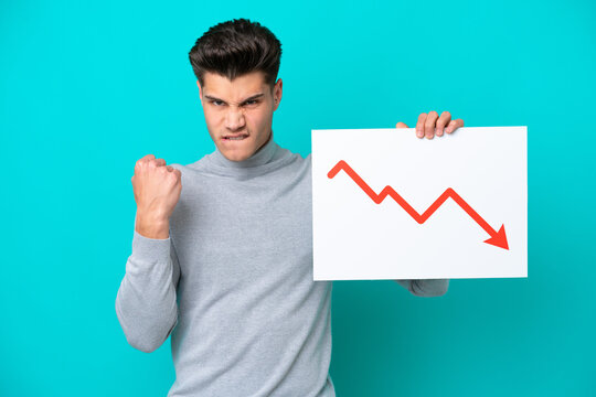 Young handsome caucasian man isolated on blue bakcground holding a sign with a decreasing statistics arrow symbol and angry