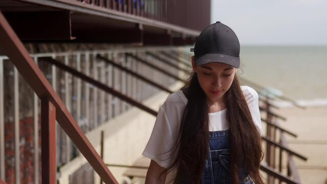 The female worker in overalls and cap goes on the construction site. Woman with long brune haire coming near a building under construction by the sea.