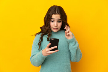 Little caucasian girl isolated on yellow background using mobile phone with fingers crossing