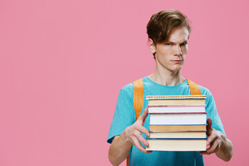 funny, red-haired student in a blue T-shirt and with an orange backpack on his back, holds a heavy stack of books in his hands and looks at the camera with surprise. Horizontal photo with empty space