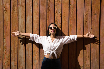 Young and beautiful woman, South American, with white knotted shirt, jeans and sunglasses with open arms on a wooden wall, in the sun. Concept beauty, fashion, vacation, travel.