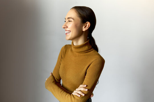 Laughing youthful girl of 20s with perfect sense of humor in stylish turtleneck standing over white background with crossed arms, laughing at funny joke, enjoying leisure time. People and emotions