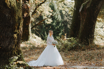 Stylish luxurious blonde bride in a white wedding dress against the backdrop of a summer sunny ancient forest.
