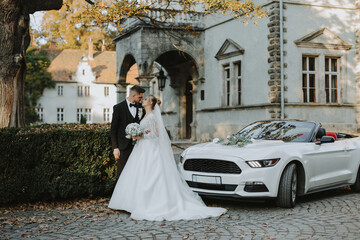 Obraz premium Stylish couple Groom and Bride are posing near the White expensive car against the backdrop of an ancient castle at sunset. The bride is holding a bouquet in her hands.
