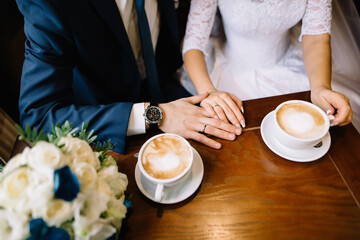A couple of newlyweds sits in a cafe and drinks coffee. Wedding gold rings on the hands. The groom gently holds the bride's hand, on the table a stylish bouquet of roses in blue and white colors
