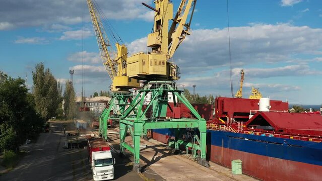 Grain transshipment from container to ship cargo holds with dual scoop clamshell grabs on gantry crane in sea port. Wheat shipment from dump trucks with water transport. Commodities transportation.