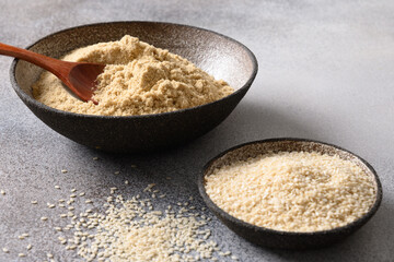 Sesame flour and white sesame seeds in bowls on gray background for cooking gluten-free and low carbohydrate dessert. Close up.