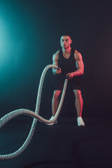 Athletic looking bodybulder work out with battle rope on dark studio background with smoke. Strength and motivation