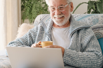 Happy elderly 70s man seated on sofa browsing by laptop holding a coffee cup, older generation and...
