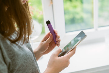  girl holds and smokes a purple disposable electronic cigarette by the window. Modern communication on the Internet