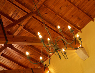 Below view of an old fashioned chandelier with candle lightbulbs hanging from sturdy wooden roof ceiling inside. Architecture beam detail, interior design, brass, antique light fixture in empty room