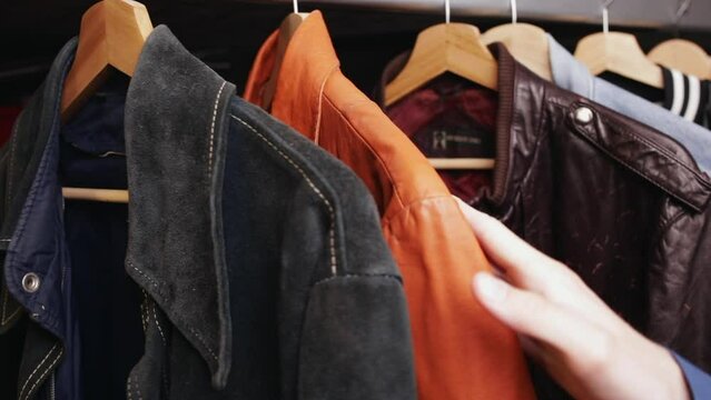 Man choosing clothing in a second hand store. Various vintage suede leather and jeans jackets hang on clothing rack. Thrifting and sustainability in clothing concept