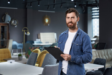 Professional sales assistant in denim shirt standing with dark folder in modern furniture store....