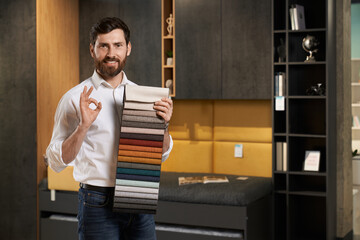 Professional male expert showing okay sign, while presenting variety samples of upholstery fabric...