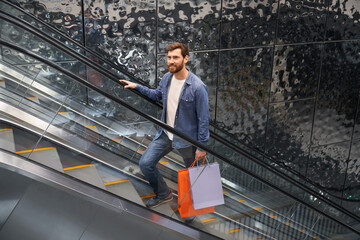 Handsome bearded male holding shopping bags, while lifting up on escalator of shopping center. View...