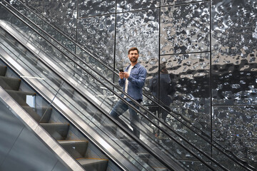 Smiling bearded male using mobile phone, while lifting on escalator. Side view of happy man standing on moving staircase, while looking away, with grey glossy tile on background. Concept of lifestyle.