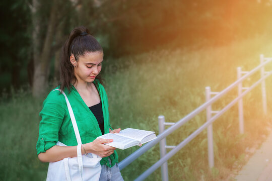 Young student girl reading book leaning to metal railing at urban park. Studying concept.