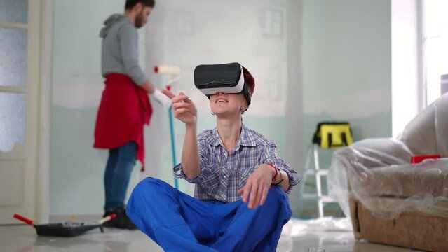 Smiling woman in VR headset gesturing examining virtual design as man painting wall at background with paint roller. Portrait of happy confident Caucasian wife indoors in new home with husband