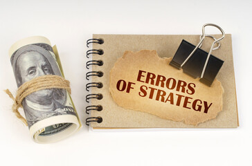 On a white surface are twisted dollars, a notebook and a cardboard sign with the inscription - Errors of strategy