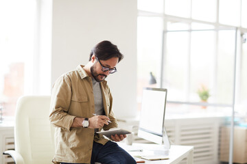 Busy handsome young Asian man in shirt sitting on table and checking website information on tablet