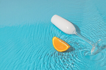 Trendy sunny scene with a glass of champagne and a slice of orange on a water surface. Summer travel concept. Minimal beach party idea.
