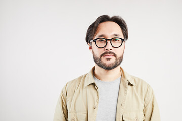 Portrait of serious modern young Asian guy in casual shirt and glasses standing against white background