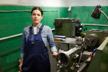 Portrait of serious middle-aged woman in overall with hanging safety goggles standing at manual lathe in industry workshop