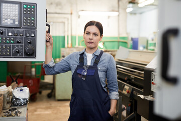 Serious confident brunette lady in overall working at factory standing by cnc lathe and holding...