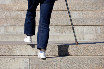 Woman wearing jeans with walking cane climbing stairs outdoor, legs in motion on the street steps. Concept for disability, limping adult, diseases of the spine, old people - obrazy, fototapety, plakaty