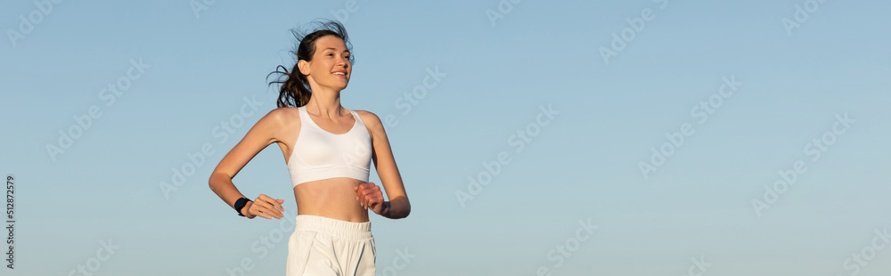 Wall mural smiling young woman in sportswear jogging against blue sky, banner.