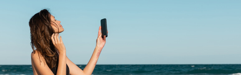 brunette young woman in swimsuit taking selfie on smartphone near sea, banner.