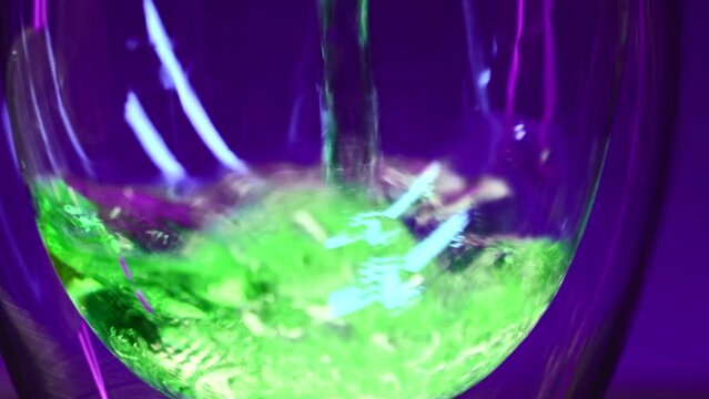 Close up of empty transparent glass being filled with green soda water. Stock clip. Wall with colorful illumination of a night bar.