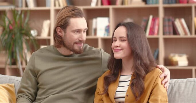 Close up on beautiful young caucasian couple man and woman sitting on sofa huggin and smiling. Happy people spending time indoors covid 19, quarantine concept.