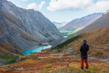 Fototapeta na wymiar Man enjoys beautiful view of blue alpine lake in colorful autumn valley. Tourist enjoying azure mountain lake scenic view from red hill among multicolor flora. Vivid autumn colors in high mountains.