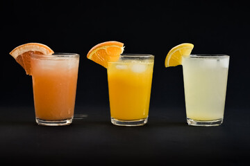 Three glasses of freshly squeezed orange juice, grape fruit juice and lemon juice lined up on a black background