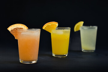 Three glasses of freshly squeezed orange juice, grape fruit juice and lemon juice lined up on a black background
