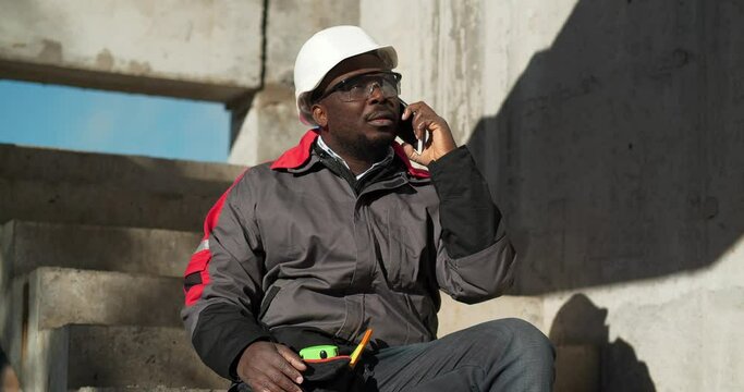 African american worker talks on smartphone. Builder in white hard hat and protective goggles sits on concrete stairs at construction site and speaks on mobile phone