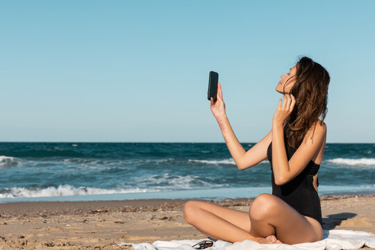 brunette young woman in swimsuit taking selfie while sitting on blanket near sea.