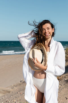cheerful young woman in white shirt and swimsuit holding straw hat on beach near sea.