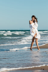 Fototapeta na wymiar full length of cheerful woman in white shirt and swimwear standing near ocean on beach.