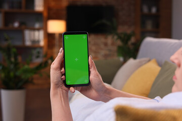 Over shoulder view of young woman holding smartphone with green mock-up screen horizontal mode.Woman lying on couch at home in evening, watching content videos blogs, news or films.Cinematic lighting.