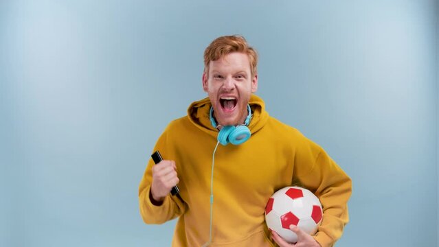Photo of funny lucky young man wear purple hoodie smiling holding ball tv remote watching football match isolated on blue background