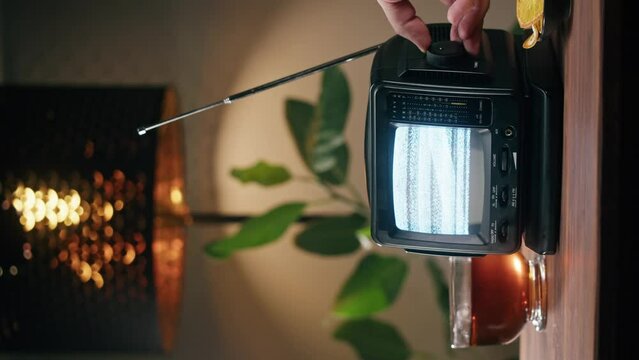Vertical video of turning on small old television with grey interference screen on home background. Close-up of vintage tv on table with plants, nostalgia. 