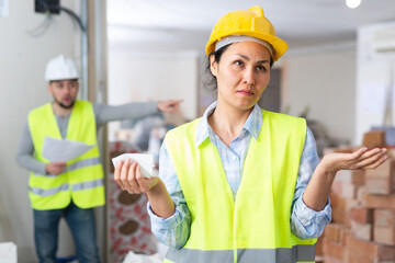Confused asian woman engineer making helpless gesture while her foreman standing in background with documents and shouting.