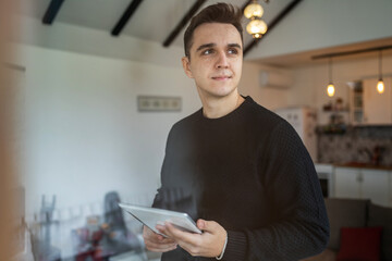 One young adult caucasian man standing indoor at home thinking and holding tablet looking to the...