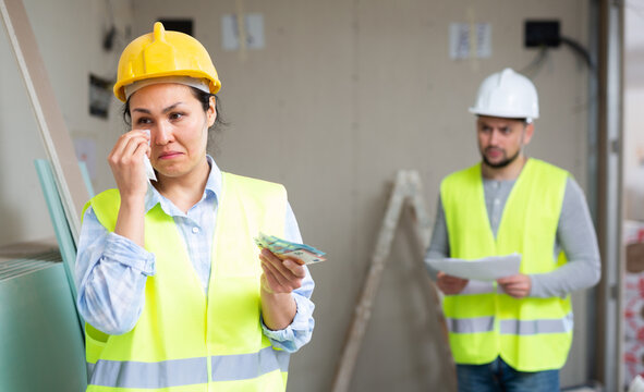 Upset Discharged Asian Female Construction Worker In Yellow Safety Vest And Hardhat Receiving Layoff Pay, Standing In Building Under Renovation With Money In Hands